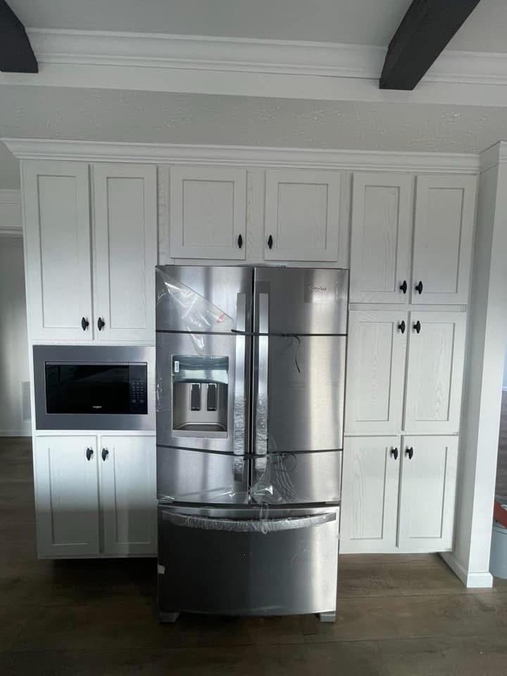 Stainless steel fridge wrapped in plastic, placed in front of white kitchen cabinets with a built-in microwave. The setting is modern and clean.