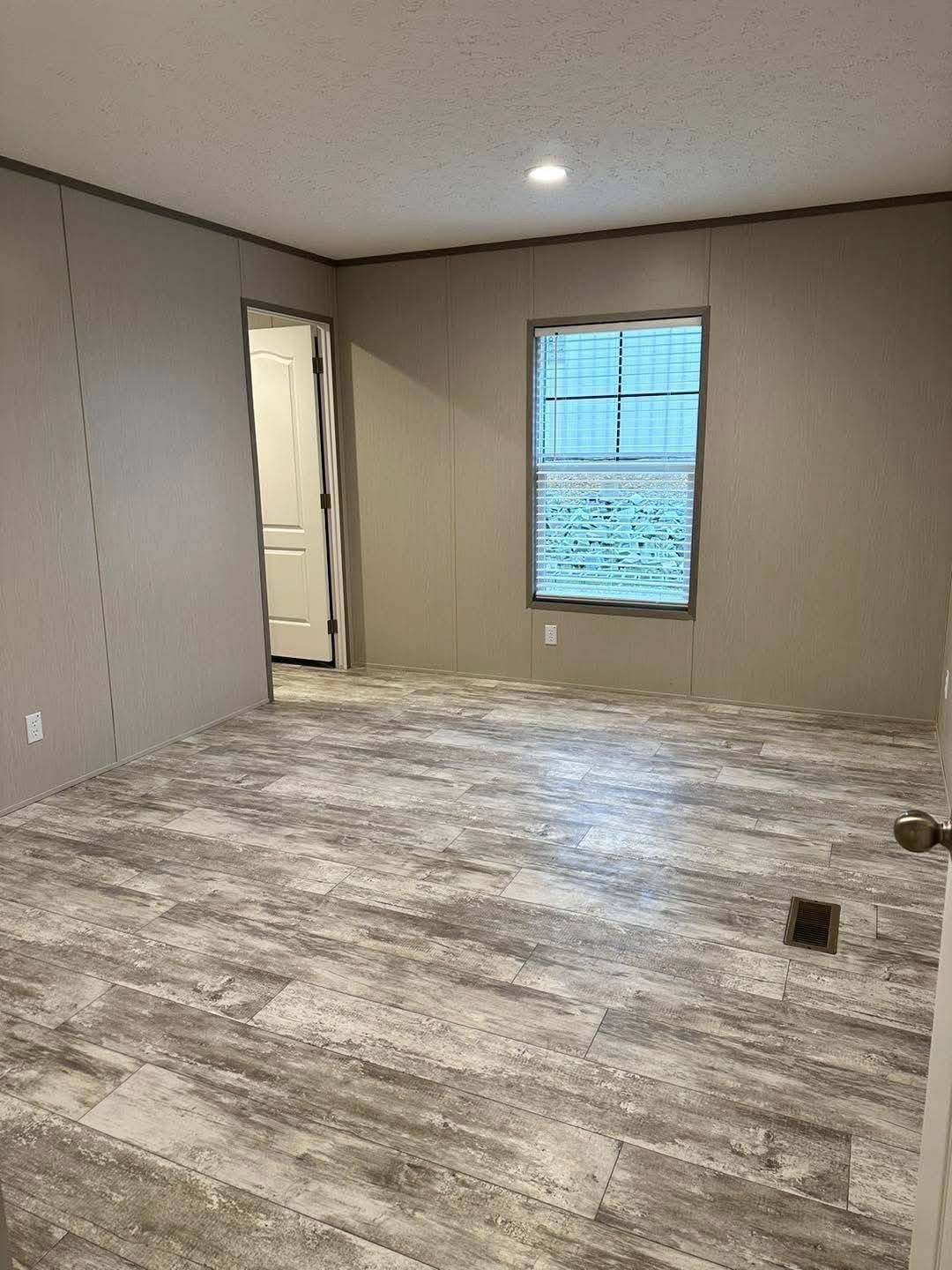 An empty room with gray wooden flooring, beige walls, and a small window on the right. The open door leads to an adjacent space, conveying simplicity.