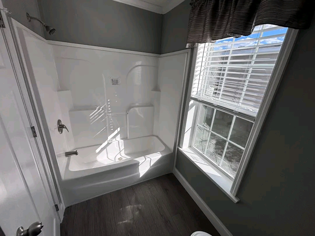Bright bathroom with a white shower-bathtub combo under a large window. Sunlight streams through blinds, casting shadows on grey walls and dark wood floor.