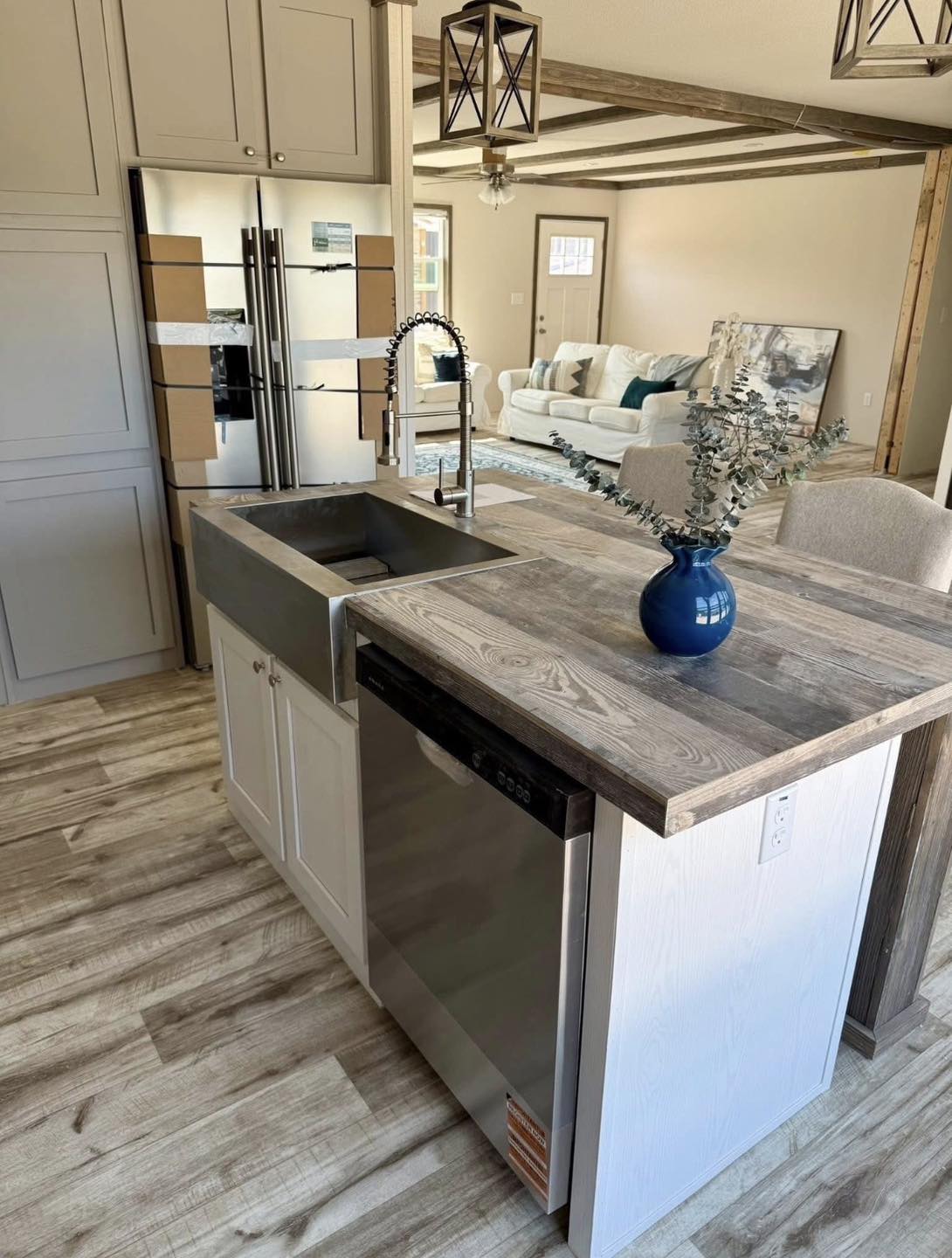 Modern kitchen interior with a wooden island featuring a stainless steel sink and dishwasher. A blue vase with greenery adds a fresh touch. Nearby, a cozy living area with a white sofa creates an inviting atmosphere.