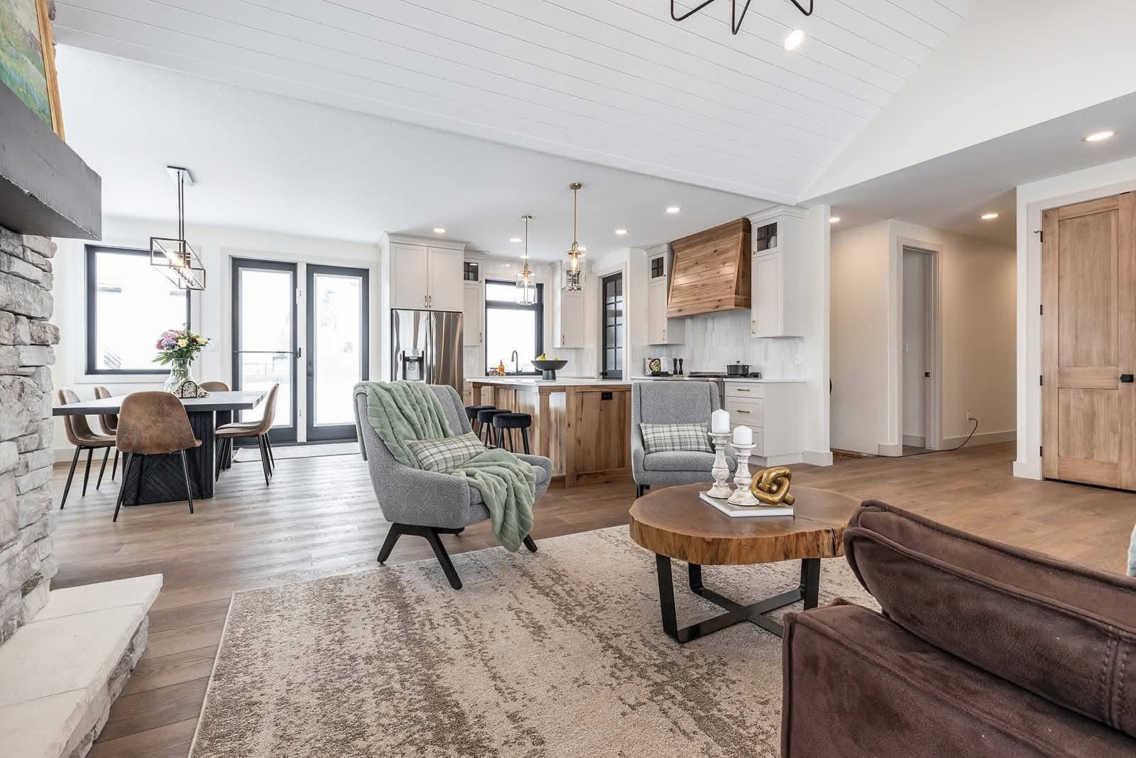 Modern open-concept living space with a gray sofa, textured rug, and wooden coffee table. Kitchen features white cabinets, and pendant lights. Airy and welcoming.