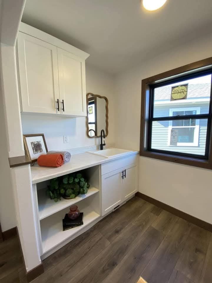 A laundry room with white cabinets, a modern black faucet, wooden shelves, and a wood-framed window. Decor includes a rolled towel and plants, conveying warmth.