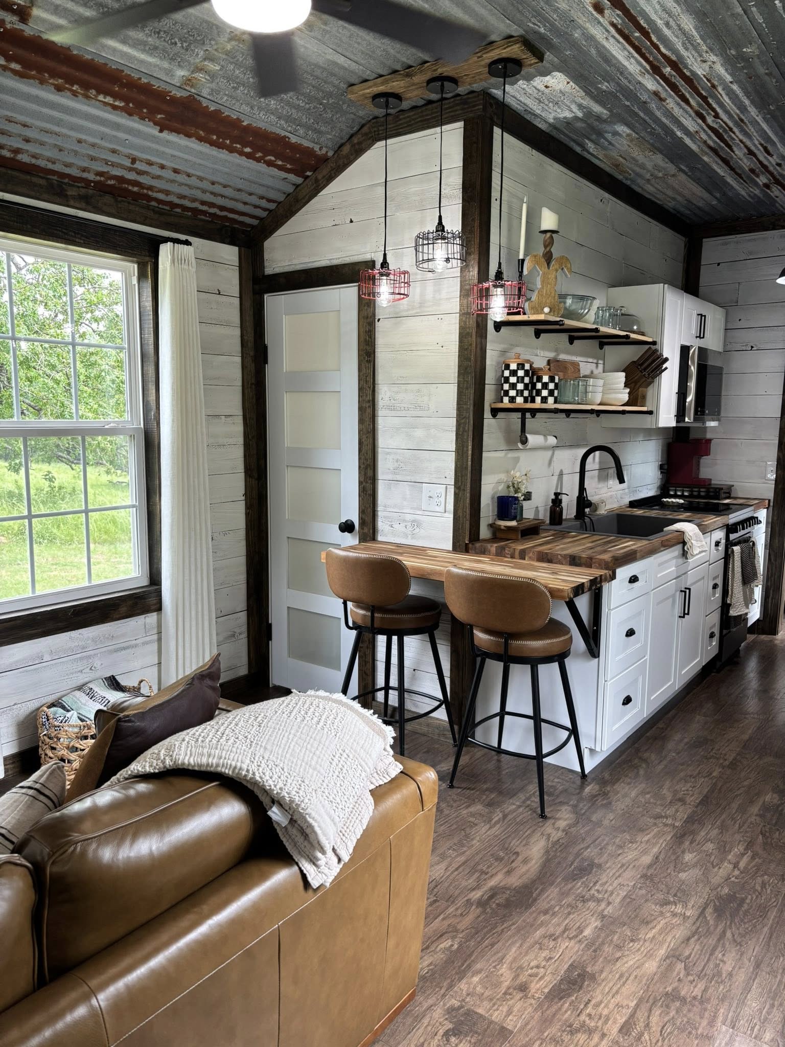 Cozy rustic kitchen and living area with wooden floors, white shiplap walls, leather sofa, wood countertop, pendant lights, and view of greenery outside.