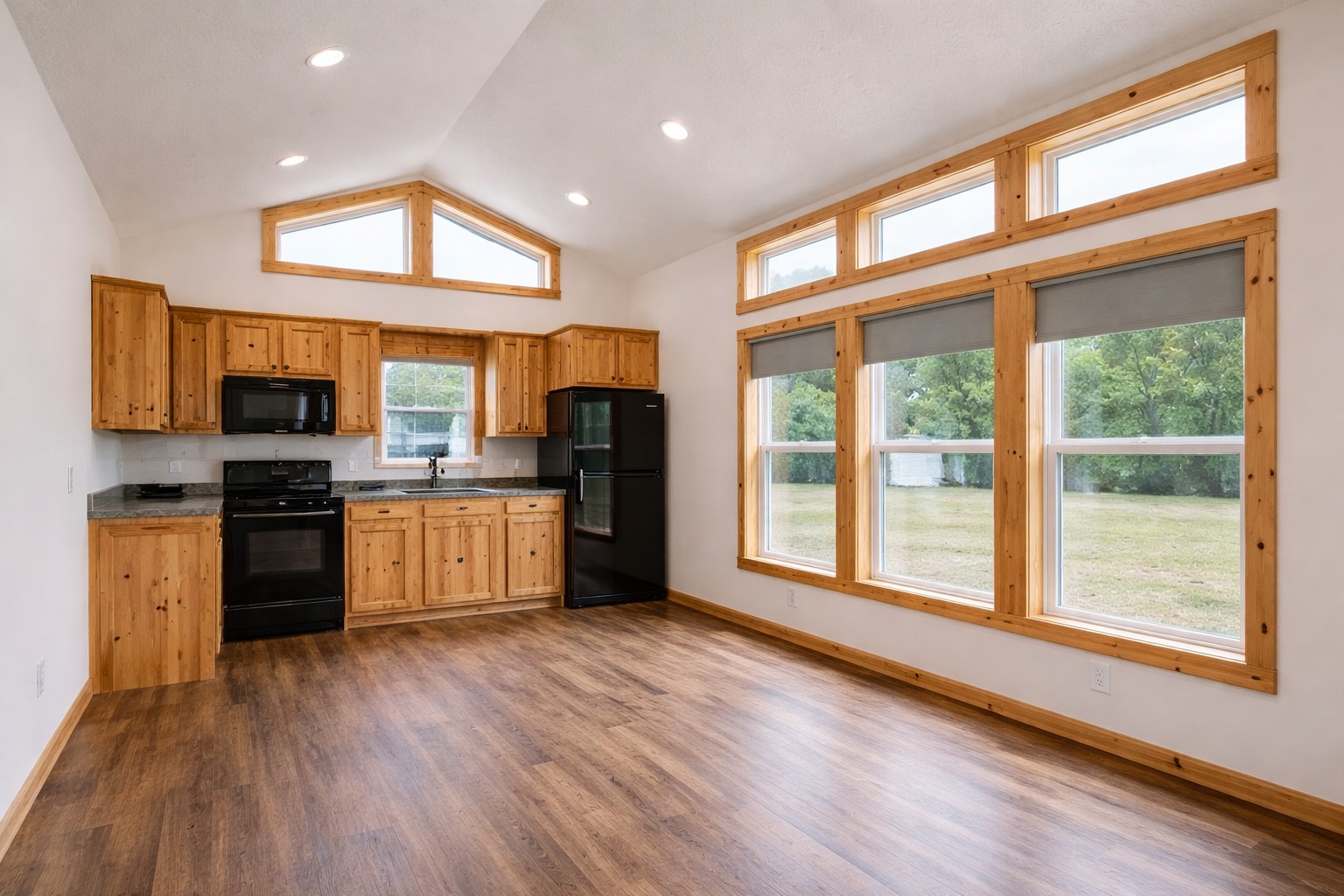 Bright kitchen with wooden floors, pine cabinets, and black appliances. Large windows with natural light and a view of green trees outside.