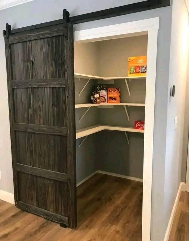 A small pantry with wooden sliding door. Inside are corner shelves holding cereal, snacks, and canned drinks. The room has wood flooring and a cozy feel.