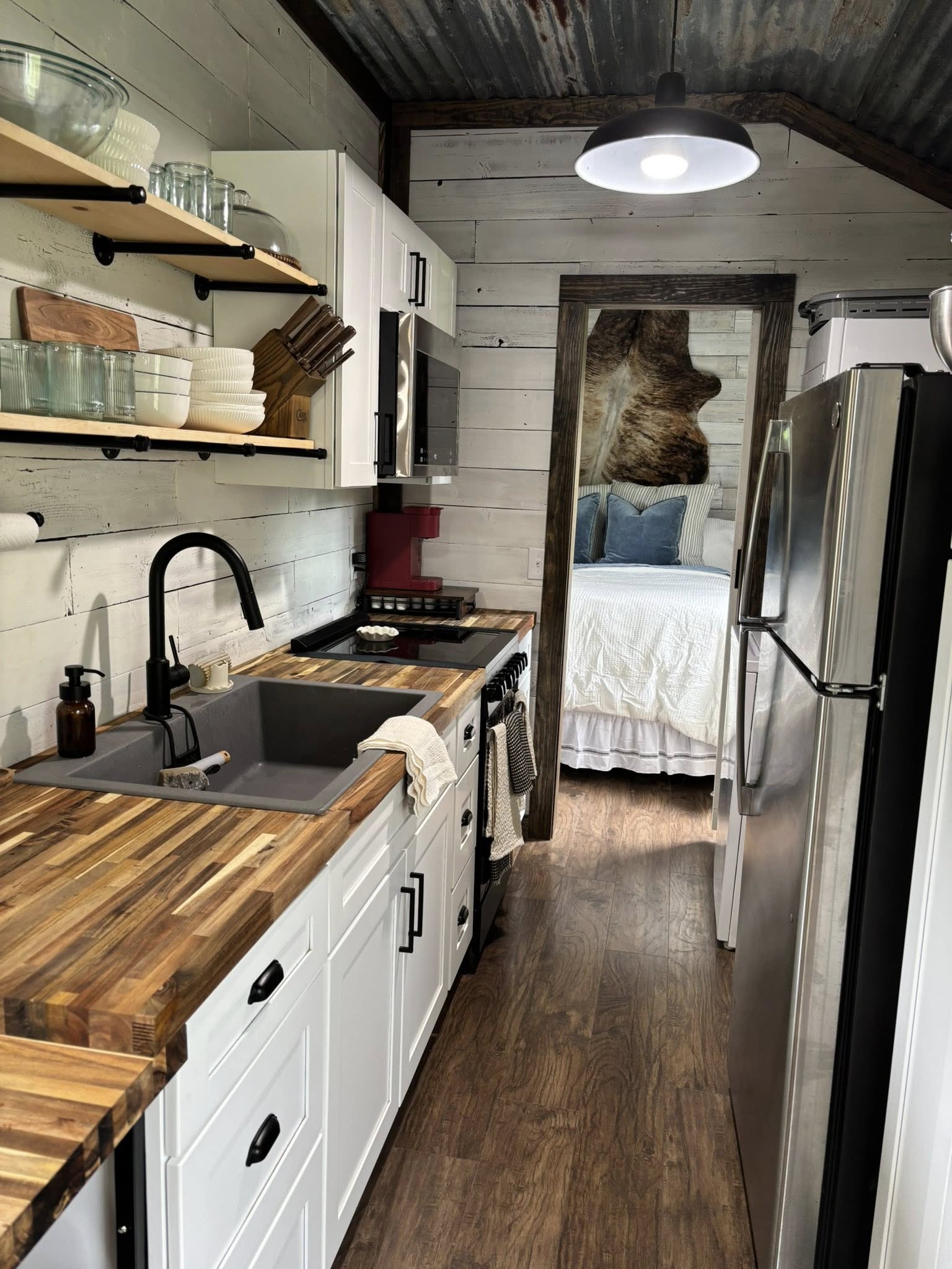 Charming galley kitchen with wood countertops, white cabinets, and open shelves. Beyond, a cozy bedroom with white bedding and accent pillows is visible.