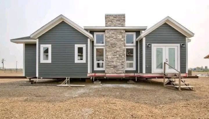 A modular home with gray siding, white trim, stone chimney, and large windows. It sits on an open, dirt area with temporary wooden steps.