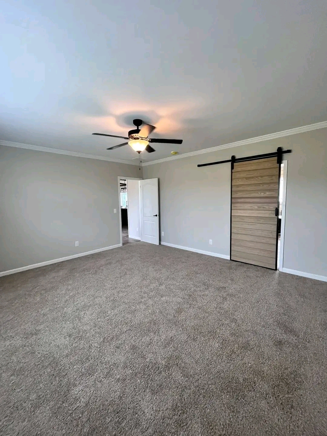 Spacious empty room with beige carpet, gray walls, and ceiling fan. Features a sliding wooden barn door and an open white door, creating a cozy feel.