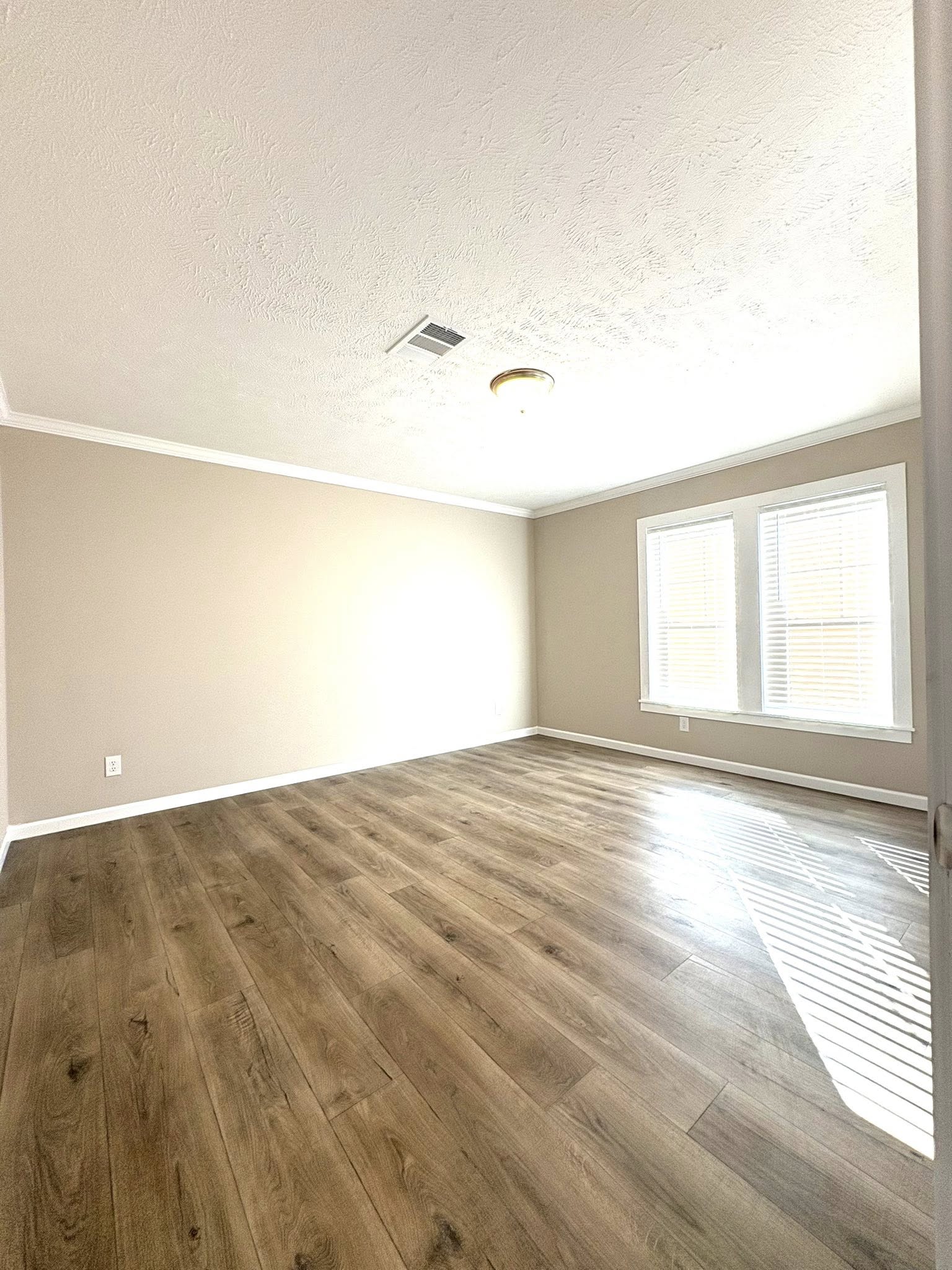 Empty room with beige walls, wood flooring, and two large windows letting in natural light. Ceiling has a simple fixture. Calm and open atmosphere.