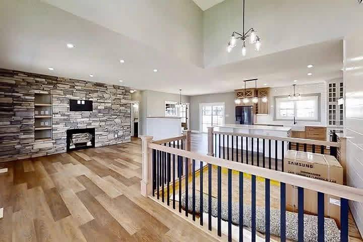 Modern open-concept living space with light wood flooring, white walls, and stone accent. Features kitchen island, pendant lights, and railing. Bright and airy.