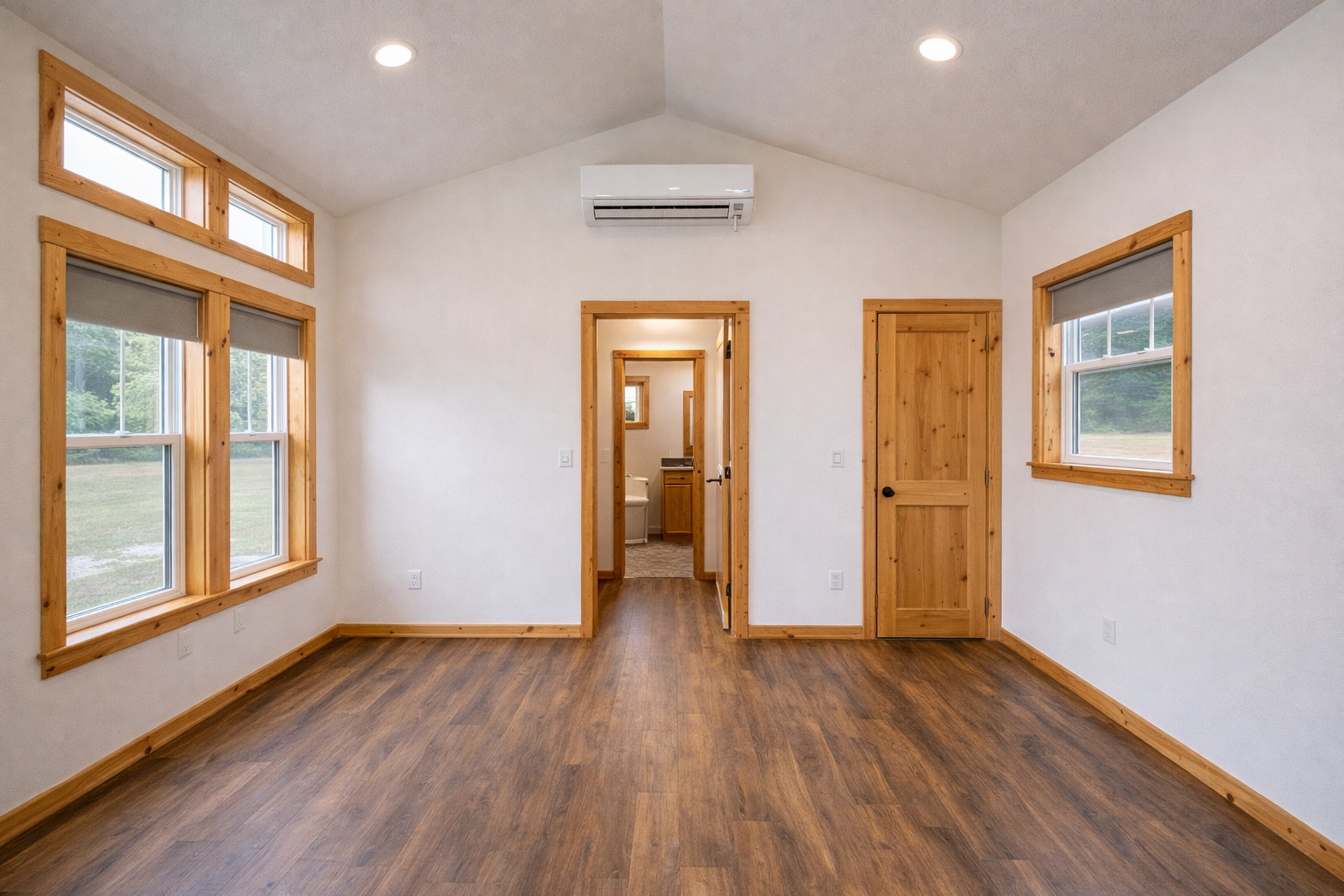 A cozy, empty room with wooden floors, white walls, and natural wood trim. Large windows let in light. A door leads to a small adjacent space.