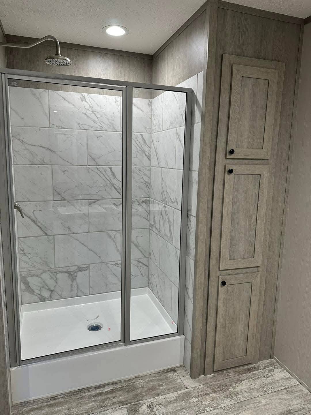 Modern bathroom with a glass-enclosed shower. The wall tiles feature a light gray marble pattern. Wooden cabinets are on the right. Bright overhead light.