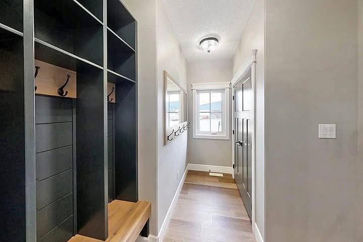 A narrow, well-lit mudroom with light gray walls features wooden cubbies and hooks on the left, a window at the end, and a door on the right.