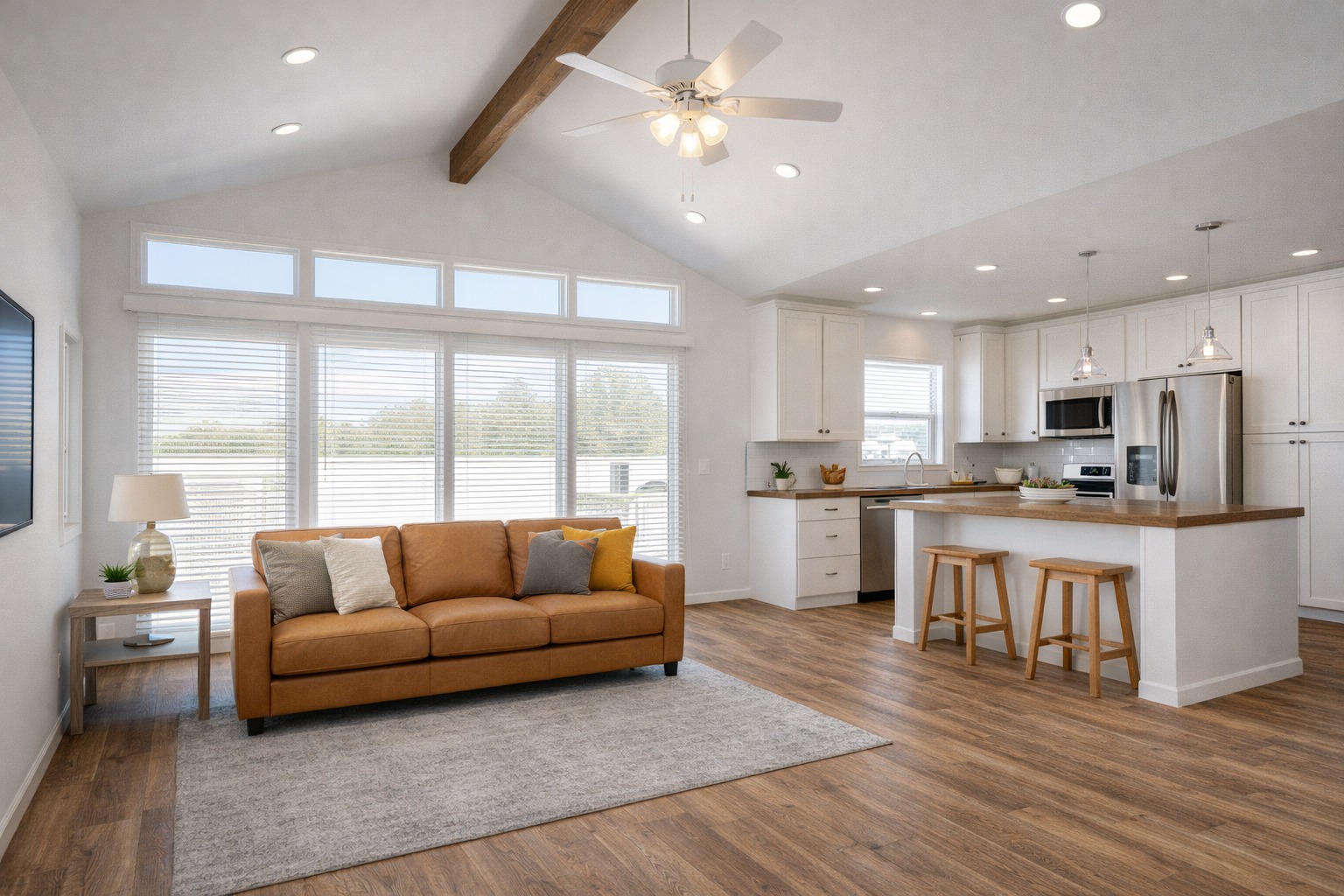 Spacious open-plan living room and kitchen with a warm ambiance. Includes a tan sofa, large windows, white cabinets, wooden floors, and bar stools.