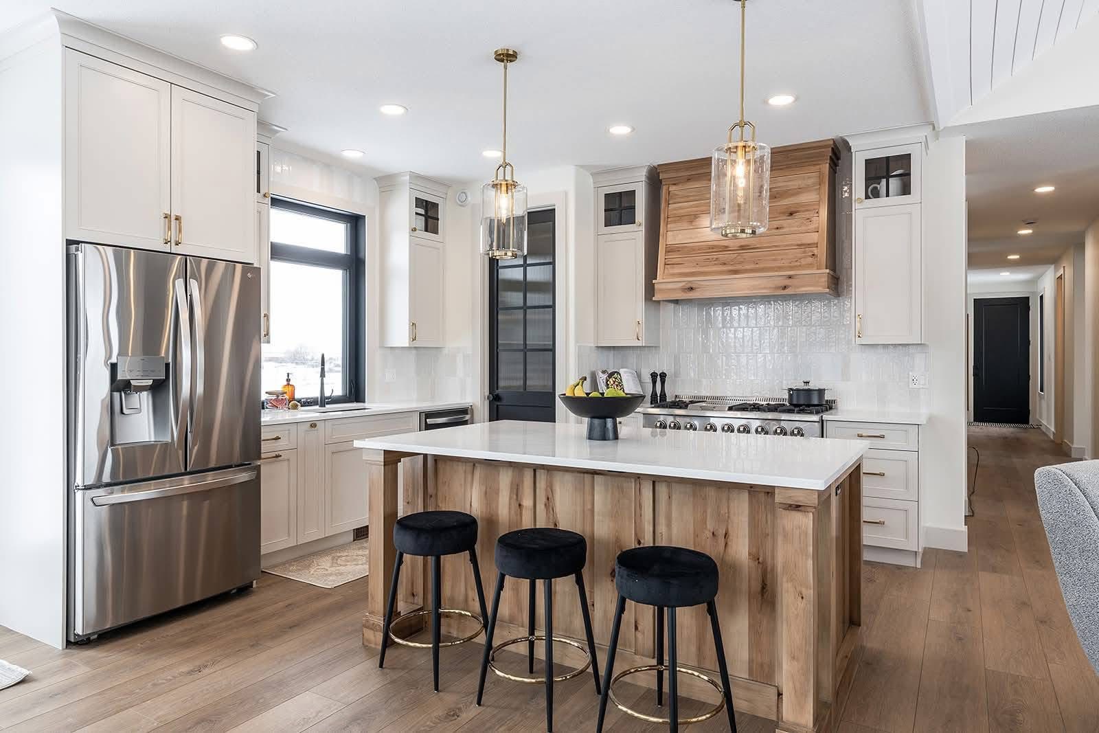 Stylish kitchen with a central island featuring wooden accents and three black stools. Stainless steel fridge, pendant lights, and light wood flooring.