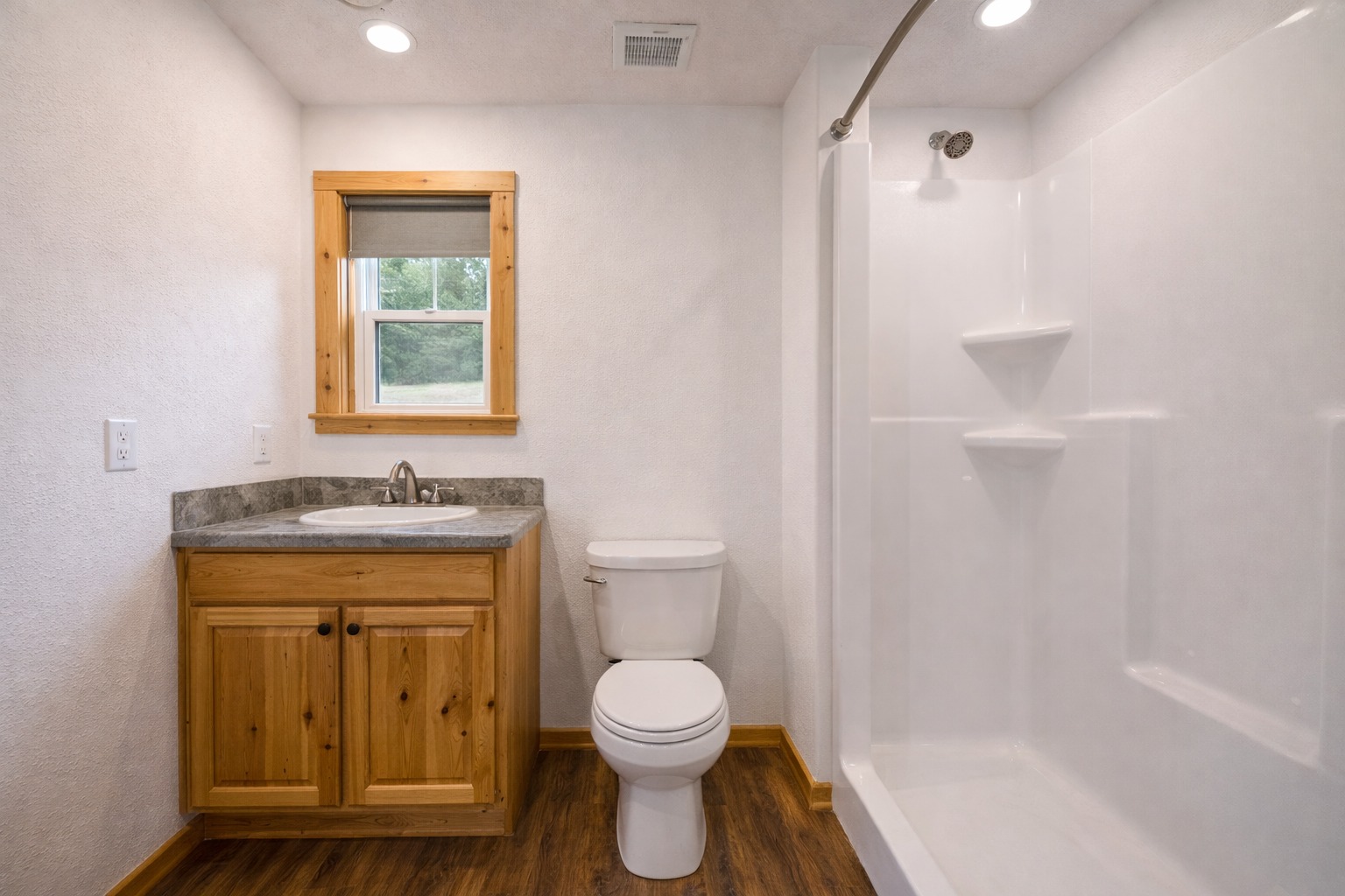 A small, modern bathroom with wooden cabinets, a ceramic sink, and a white toilet. It has a walk-in shower and a window offering natural light.