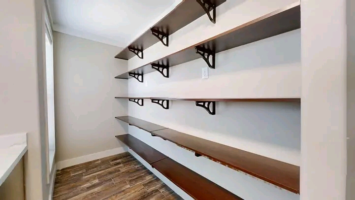 Room with four dark wooden shelves mounted on a white wall using black brackets, adjacent to a window. Floor has wood-like tiles, creating a minimalist and organized feel.