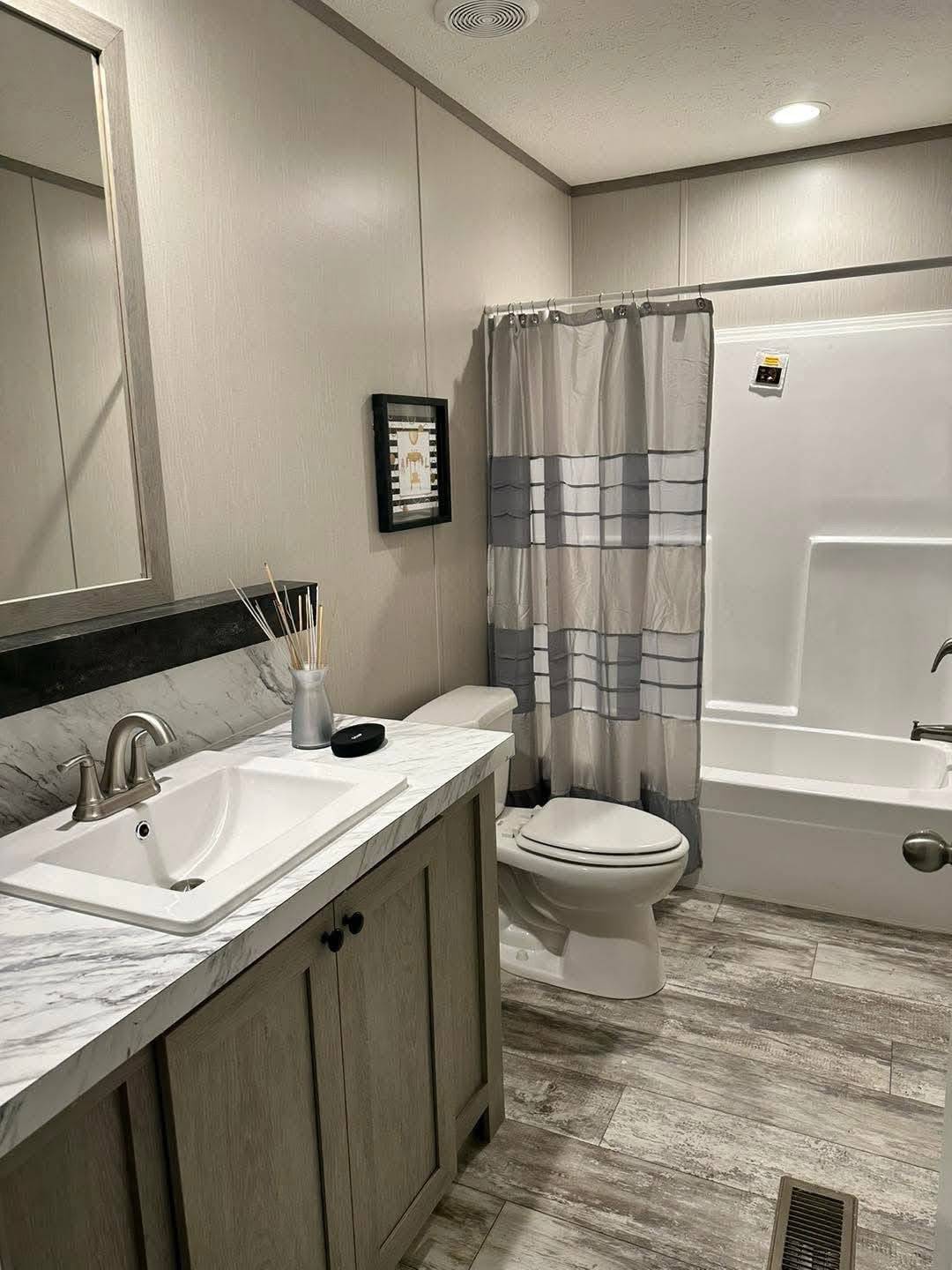 Modern bathroom with a marble countertop sink, gray wood cabinet, striped shower curtain, and wood-patterned tile floor. Neutral tones create a calm atmosphere.