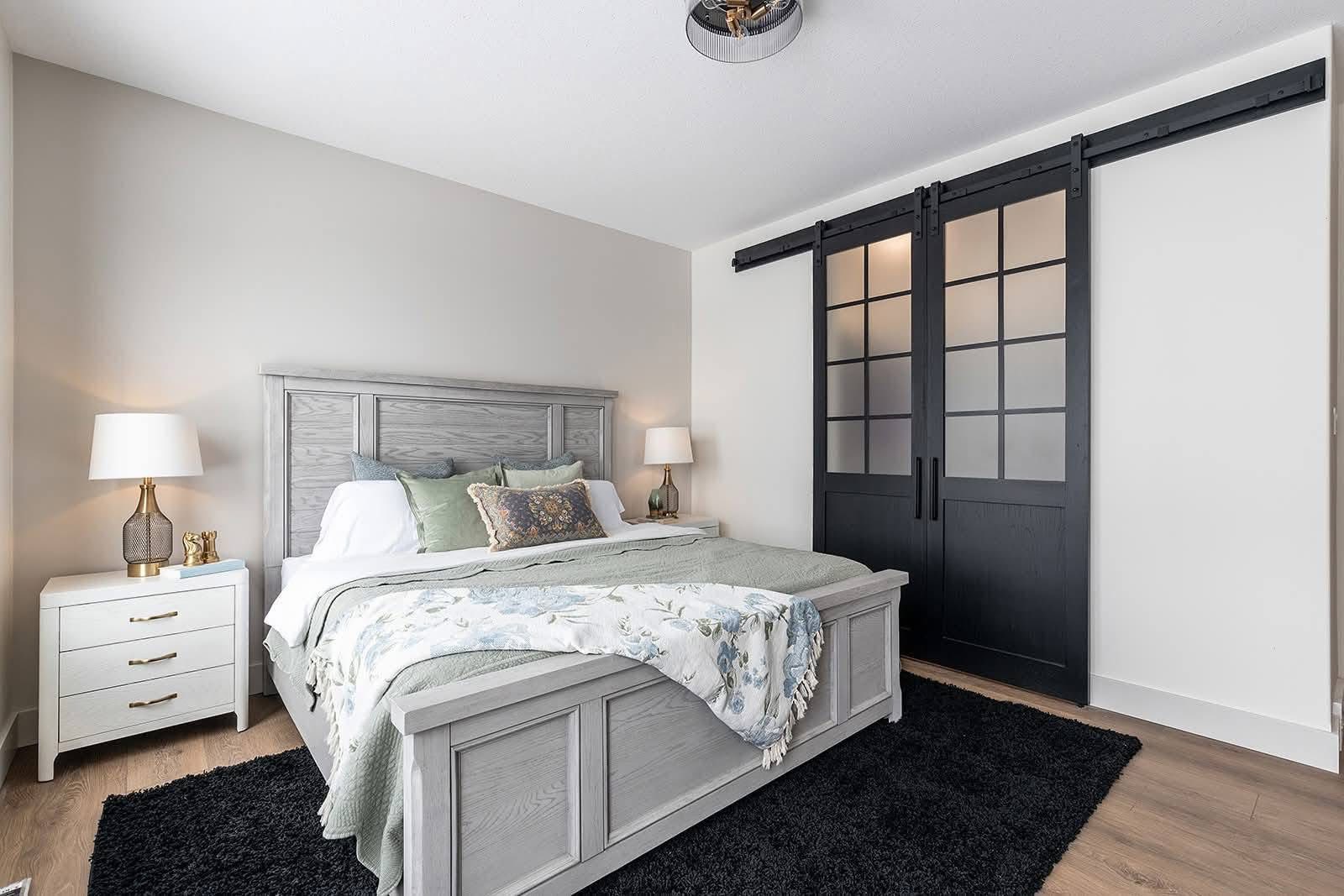 A serene bedroom with a gray wooden bed, soft green and floral bedding, and matching pillows. White nightstands with lamps flank the bed. Black sliding barn doors add contrast, and light wood flooring with a black rug completes the cozy, modern look.