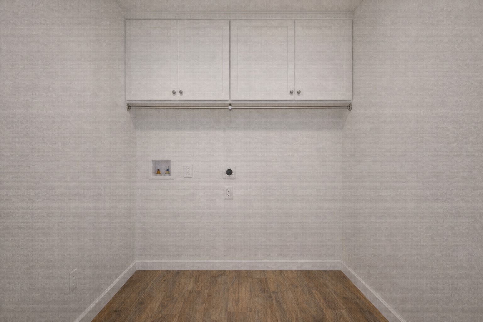 Small, empty laundry room with white walls and wood flooring. White cabinets above a laundry hookup area create a clean, minimalist feel.