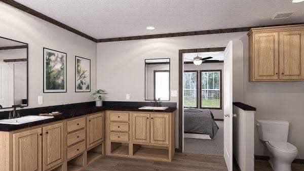Spacious bathroom features light wood cabinets, two sinks, and framed art on the walls. An open door reveals a bedroom, creating a calm, welcoming ambiance.