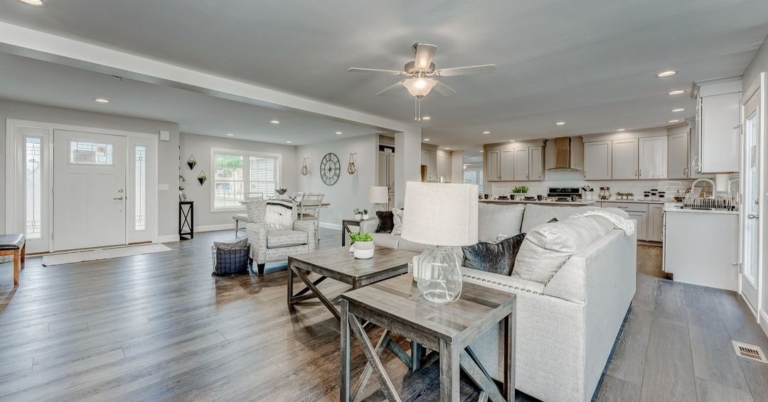 Open-concept living room and kitchen with a cozy, modern feel. Gray sofas, wooden tables, and sleek kitchen cabinets create a welcoming atmosphere.