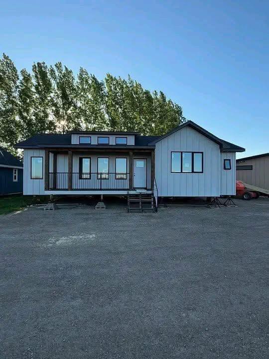 Single-story prefabricated house on a gravel lot, with large windows, steps leading to a porch, and trees in the background against a clear sky.
