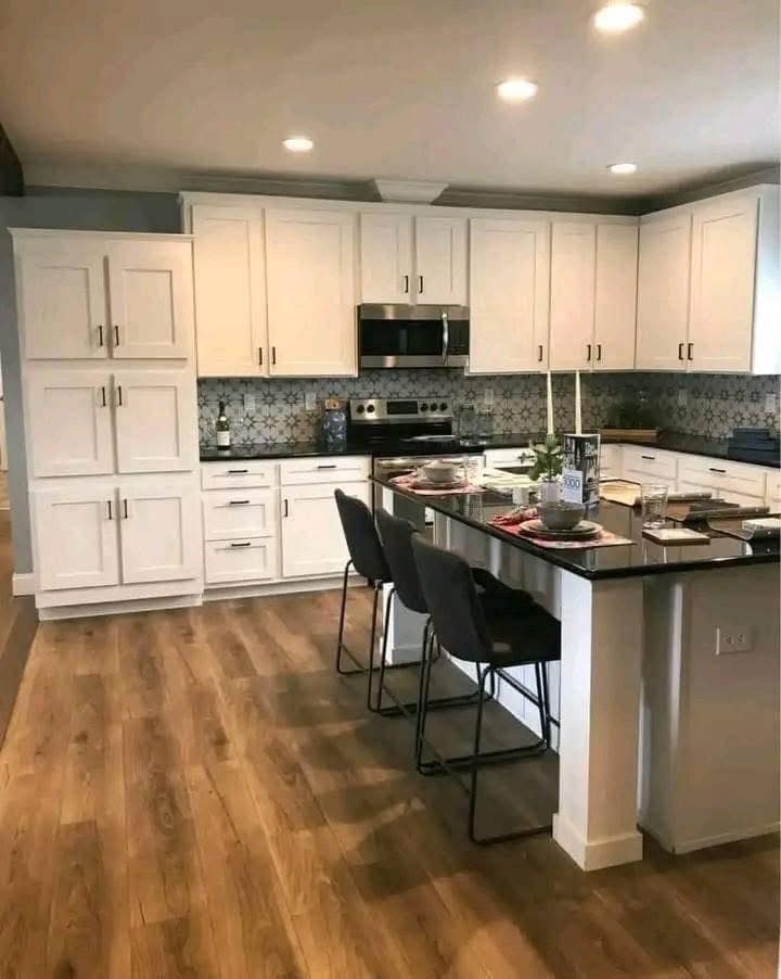 Modern kitchen with white cabinets, patterned backsplash, and stainless steel appliances. An island with gray chairs and wood flooring adds warmth.