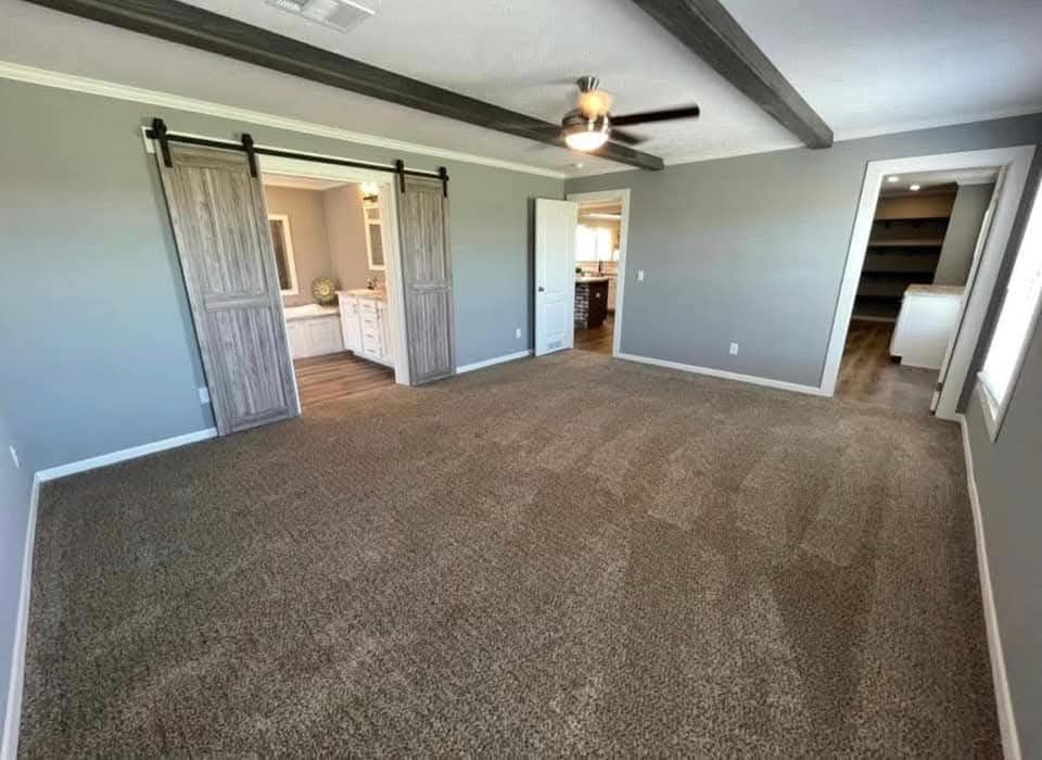 Spacious carpeted room with gray walls and ceiling beams. Features a ceiling fan, barn doors leading to a bathroom, and an open door to a closet.