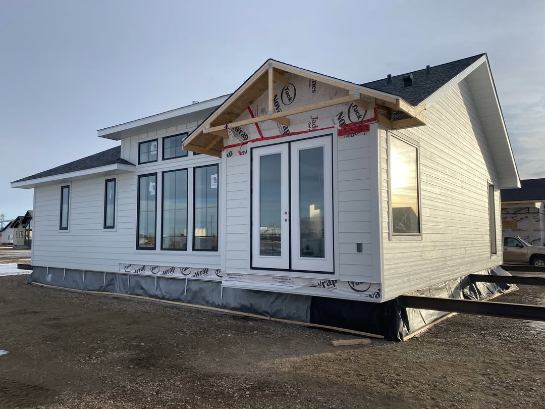 Single-story house under construction with white siding and large windows. The clear sky and sunlight suggest a calm and bright atmosphere.