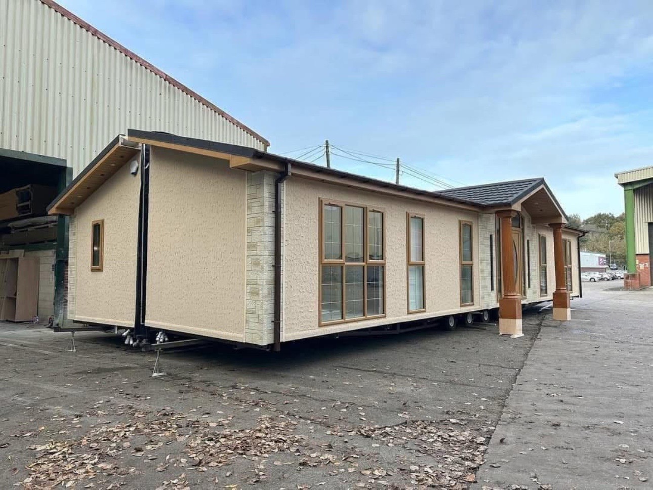 A beige mobile home with large windows and brown trim on a lot, surrounded by industrial buildings and scattered leaves, under a cloudy sky.
