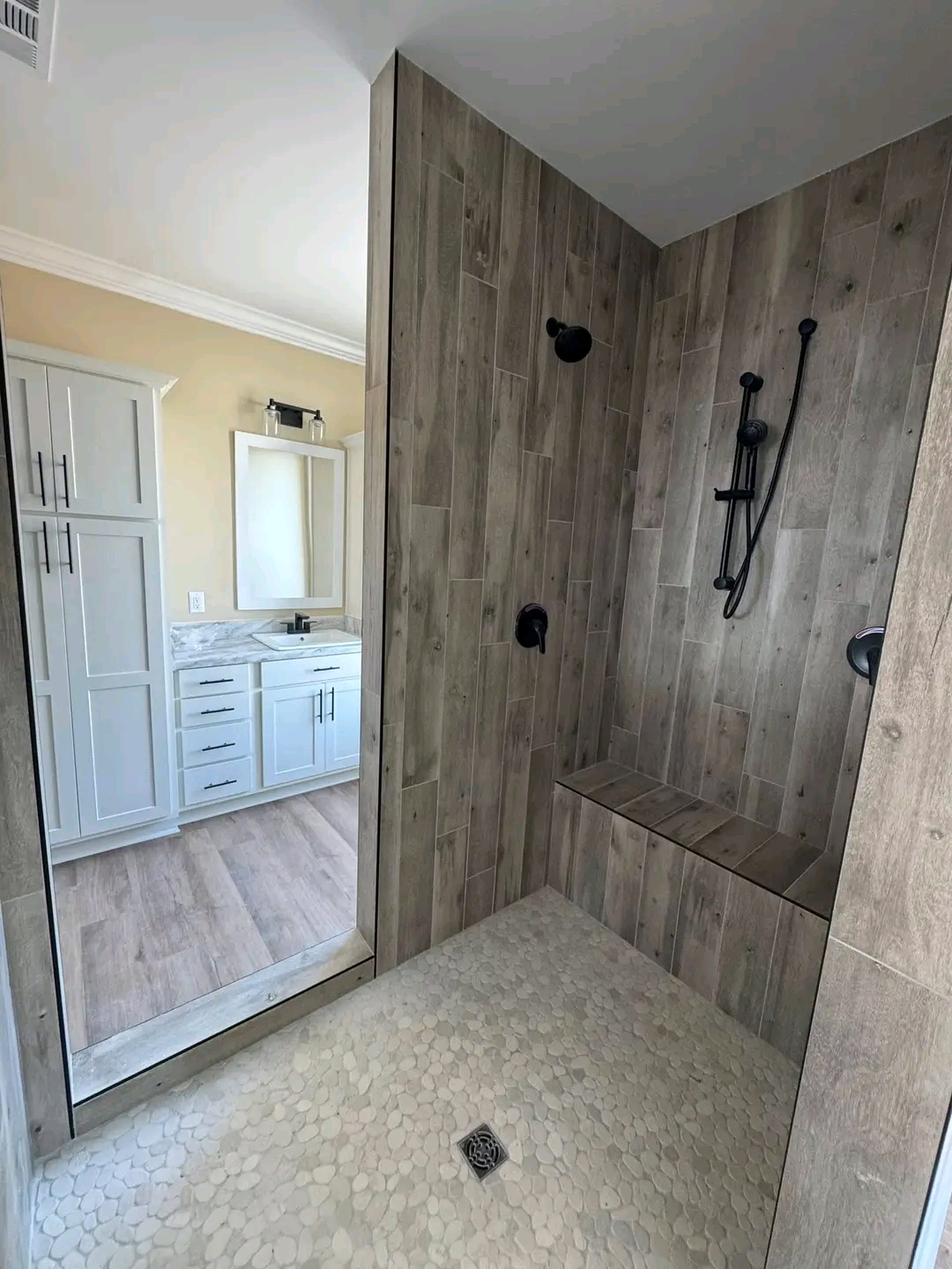 Modern bathroom featuring a walk-in shower with wood-like tiles and pebble flooring. Adjacent is a white vanity with a marble countertop and cabinet.