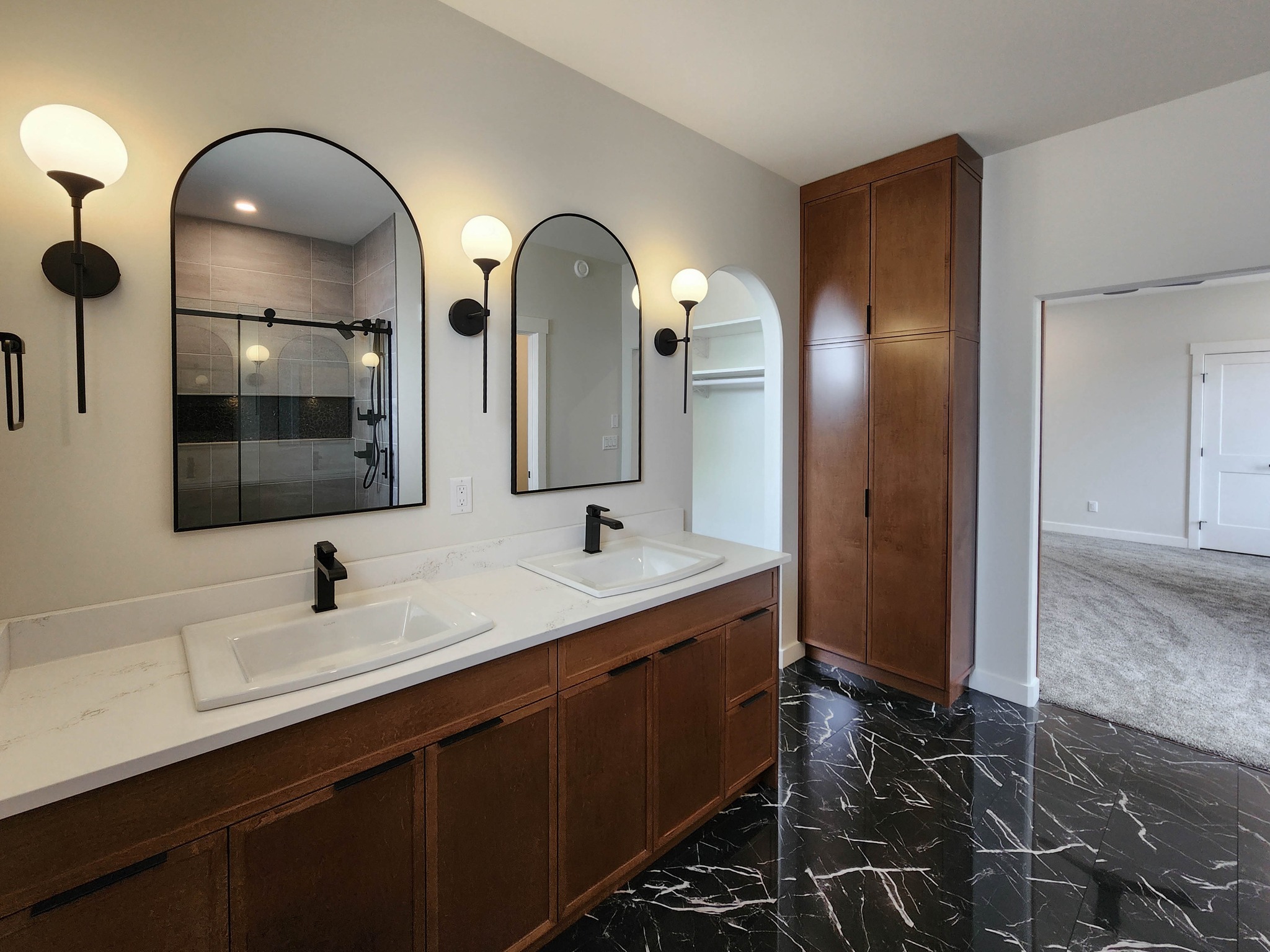 Modern bathroom with dual sinks on a wooden vanity, black fixtures, and arched mirrors. Marble black floor, warm lighting, and adjacent walk-in closet.