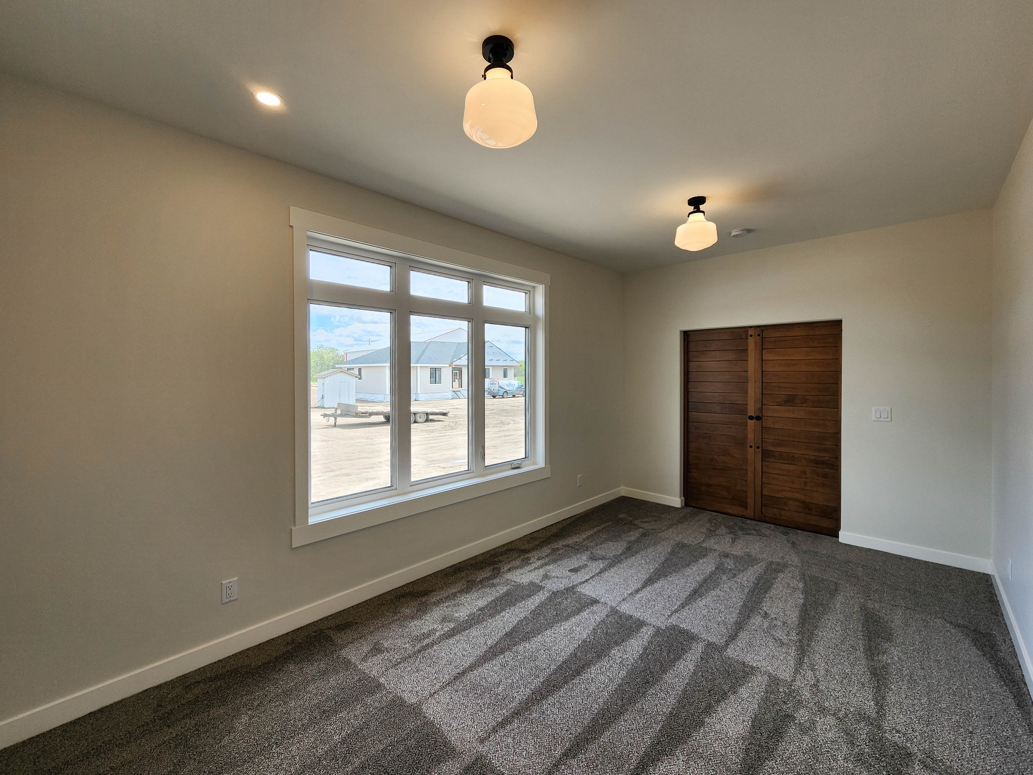 Empty room with neutral walls, large window, and carpeted floor. Wooden double doors on the right. Two ceiling lights create a calm, welcoming atmosphere.