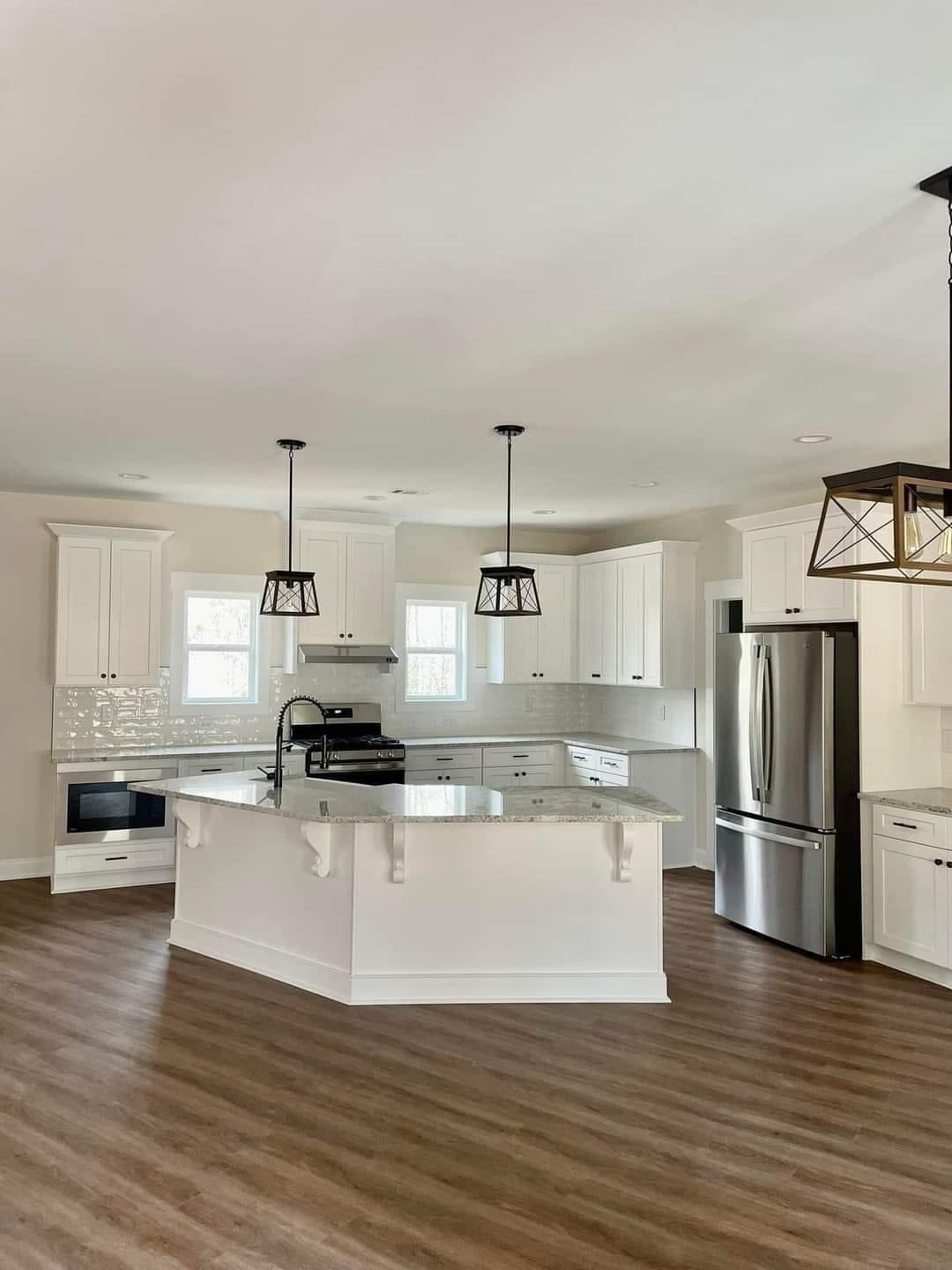 Spacious kitchen with white cabinets, granite island, stainless steel appliances, and wood flooring. Two pendant lights hang over the island. Bright and modern.