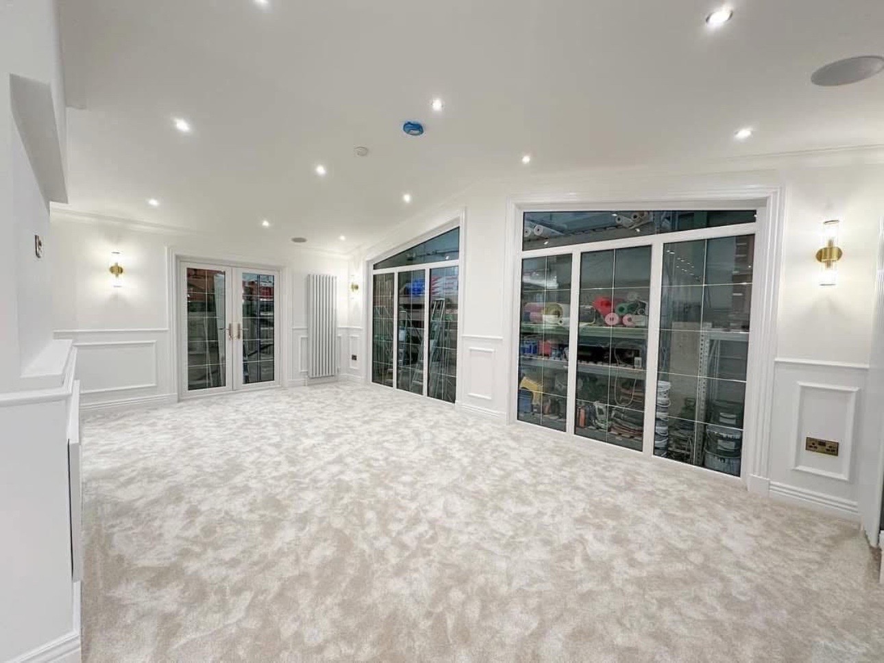 Spacious white room with plush carpet, lit by recessed lights. Large windows and glass doors reveal a storage area with shelves. Elegant, modern ambiance.