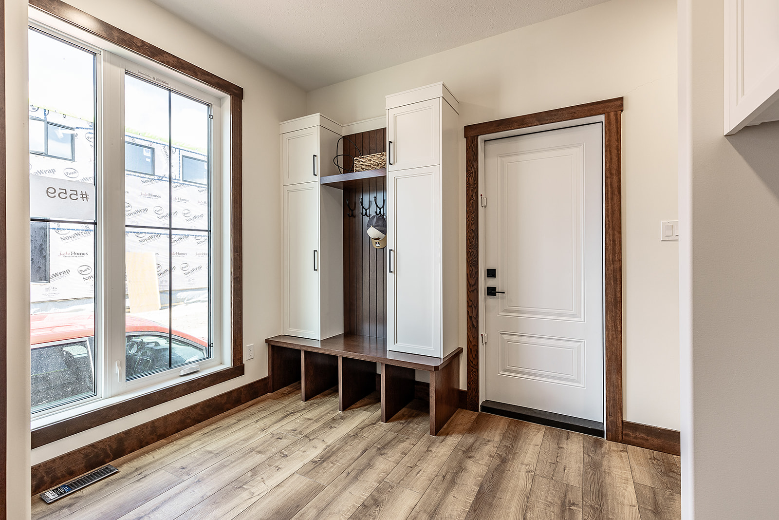 Bright entryway with wooden floor and white walls. Features a white door with dark wood trim and a custom storage bench with cabinets and hooks.