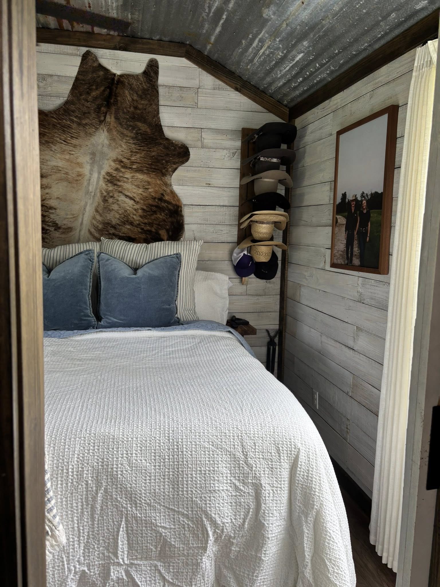 Cozy rustic bedroom with a white quilted bed, blue pillows, cowhide wall decor, and hats on a rack. A framed photo adds a personal touch.