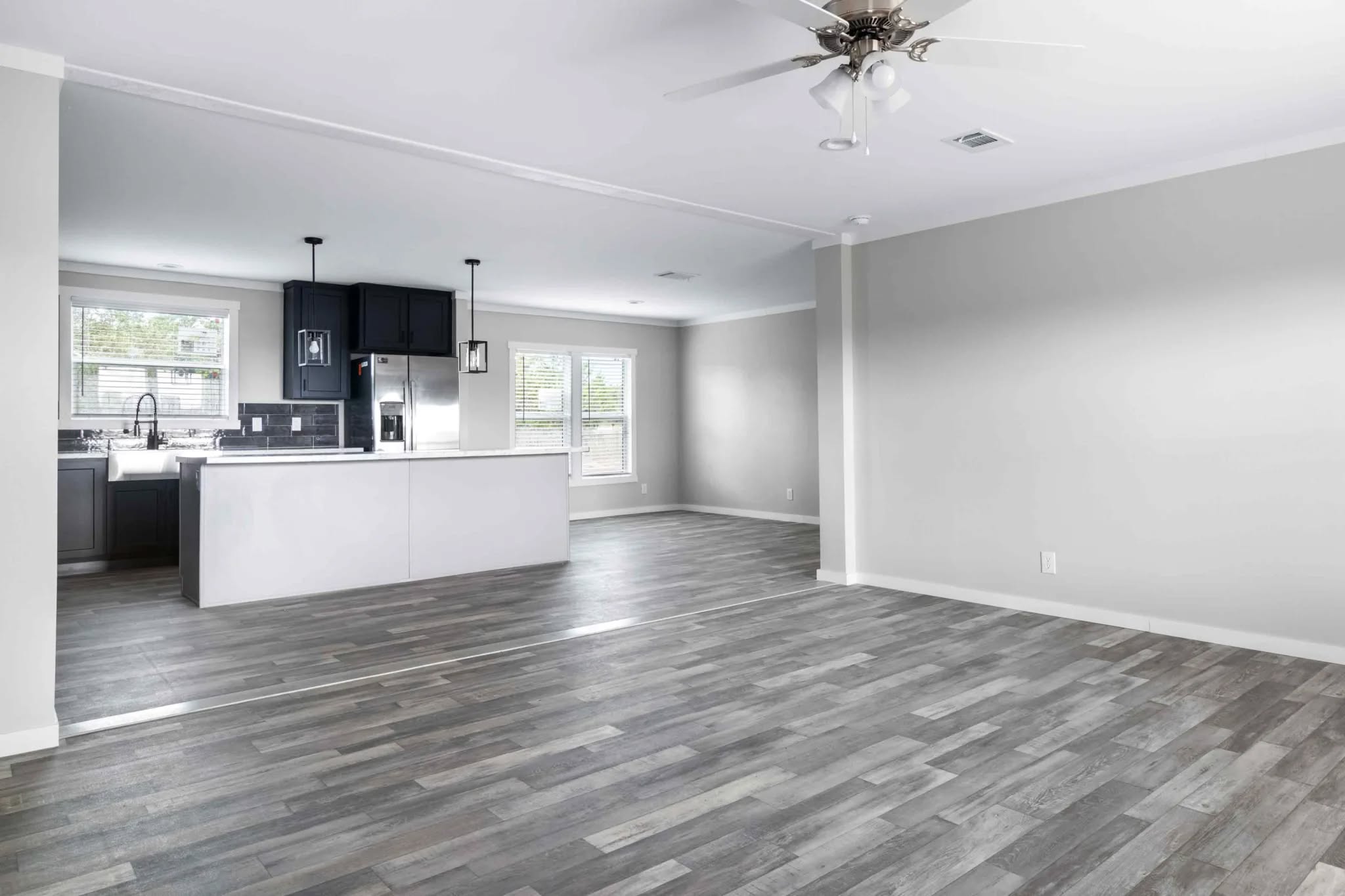 Modern open-concept space with gray wood flooring, light gray walls, and recessed ceiling lights. The kitchen features a white island, dark cabinets, and pendant lights, exuding a contemporary and airy feel.