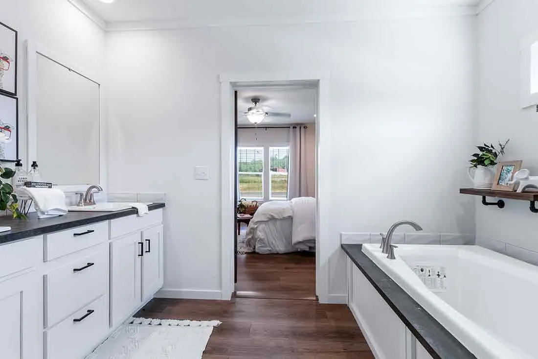 Modern bathroom with white cabinets and black countertops. A spacious bathtub is on the right, leading to a cozy bedroom with large windows.