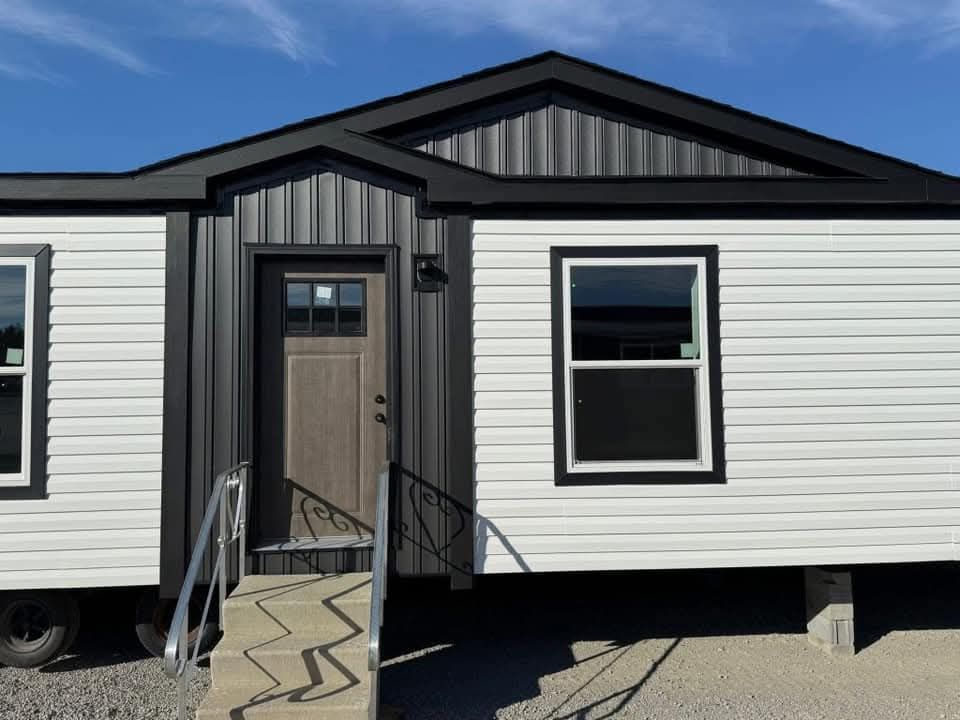 A tiny house exterior with white siding, a dark metal roof, and two windows. The wooden door has a small porch with angled metal railings. Bright, sunny day.