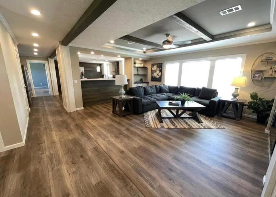 Spacious living room with wood flooring and ceiling beams. Features a gray sectional sofa, coffee table, and large windows with natural light. Cozy atmosphere.