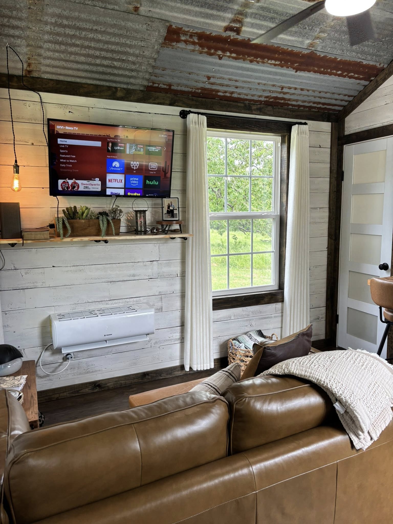 Cozy living room with leather sofa, rustic white walls, and metal ceiling. A TV and shelf with plants adorn the wall. Sunlight floods through a large window.