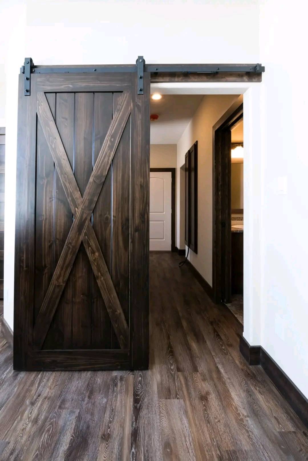 Sliding barn door with dark wood grain and X pattern, partially open to reveal a hallway with wooden flooring and white walls, creating a rustic, warm ambiance.
