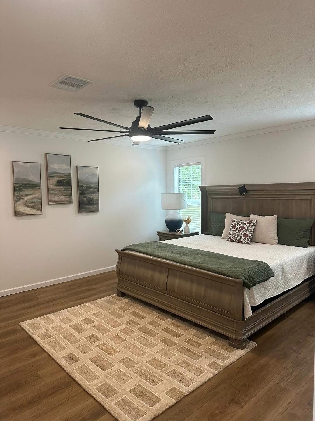 Spacious bedroom with wooden bed, green accents, and neutral bedding. A large ceiling fan, three landscape paintings, and a beige rug complete the serene setting.