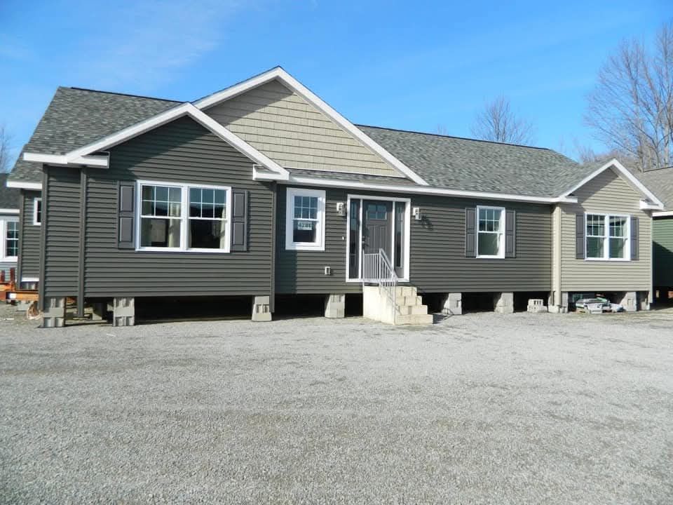 A modular home with dark green siding, white trim, and a gabled roof sits elevated on blocks. It is surrounded by a gravel lot under a clear blue sky.