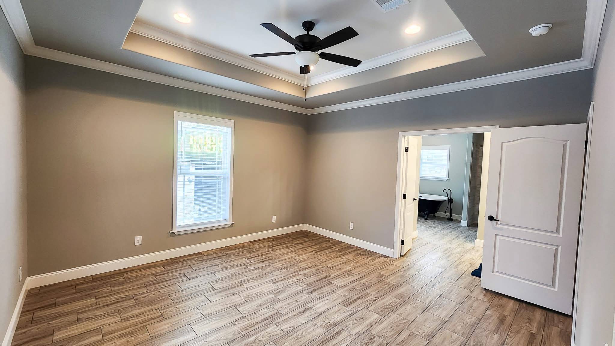 A spacious, empty room with light gray walls and wood-patterned flooring. It features a tray ceiling with a black ceiling fan and recessed lighting. A large window on the left lets in natural light, and an open door leads to an adjoining room. The atmosphere is modern and inviting.