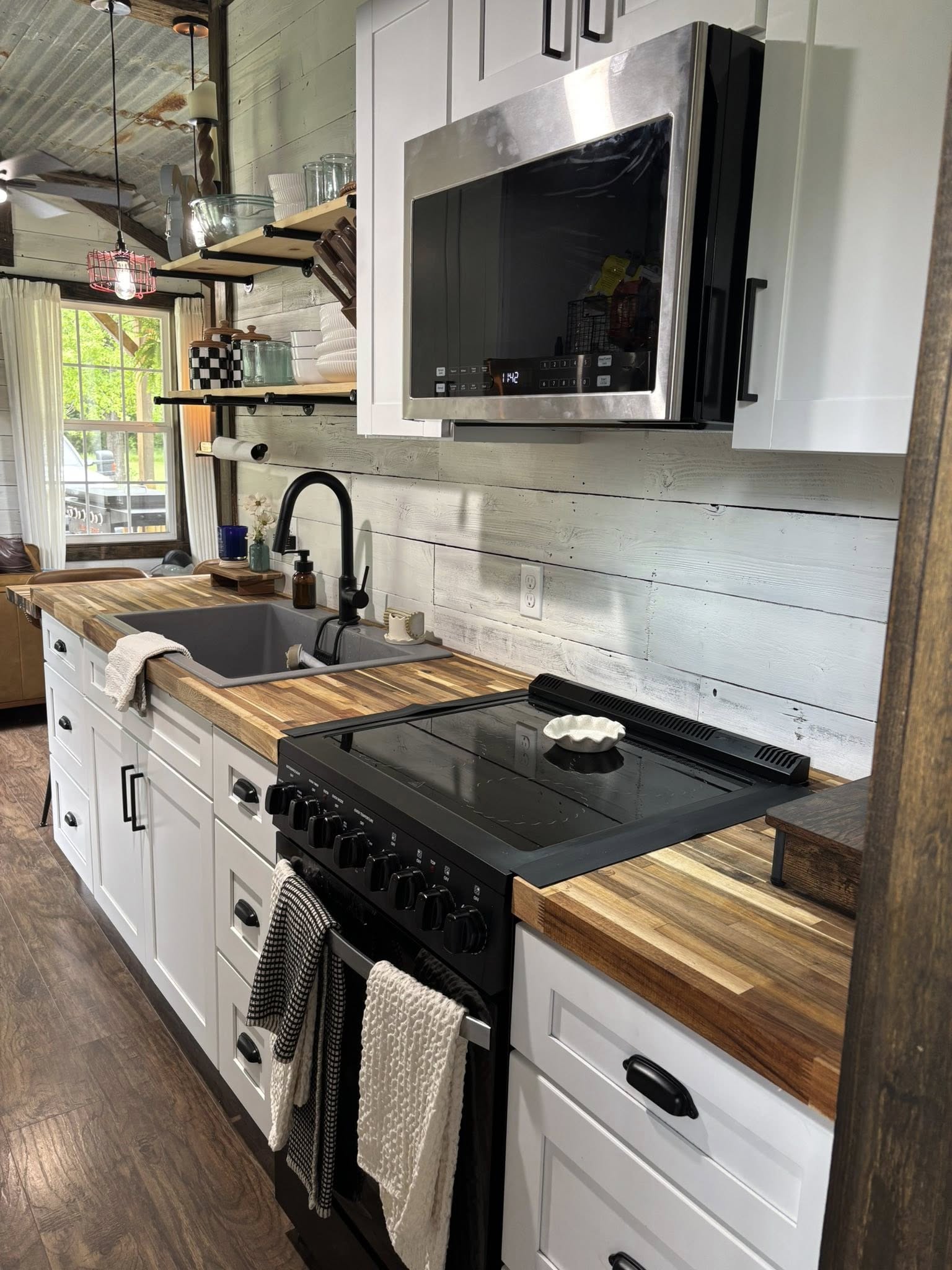 Modern kitchen with rustic charm, featuring wooden countertops, a black stove, white cabinets, open shelving, and a farmhouse sink. Bright window view.