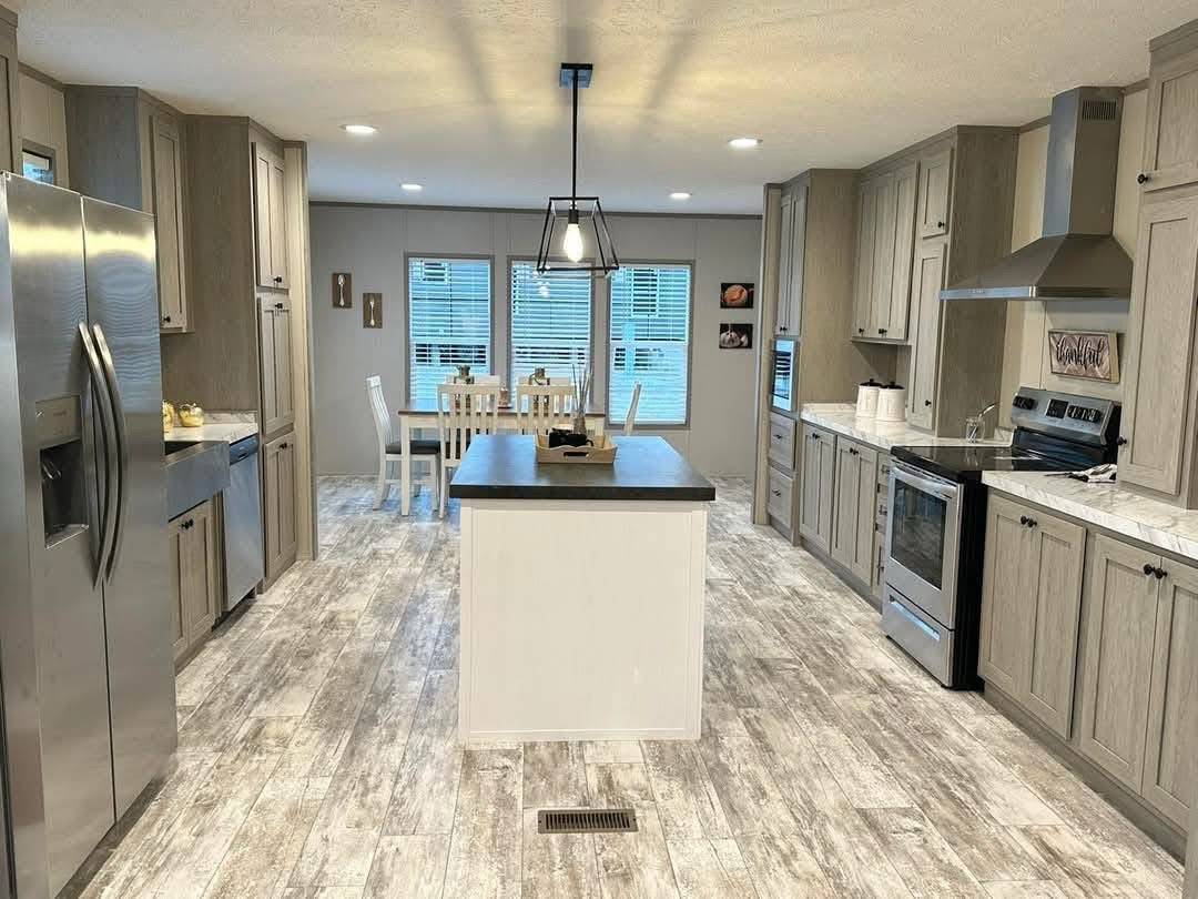 Spacious kitchen with light wood cabinets, a central island, stainless steel appliances, and rustic flooring. Cozy dining area with white chairs by large windows.
