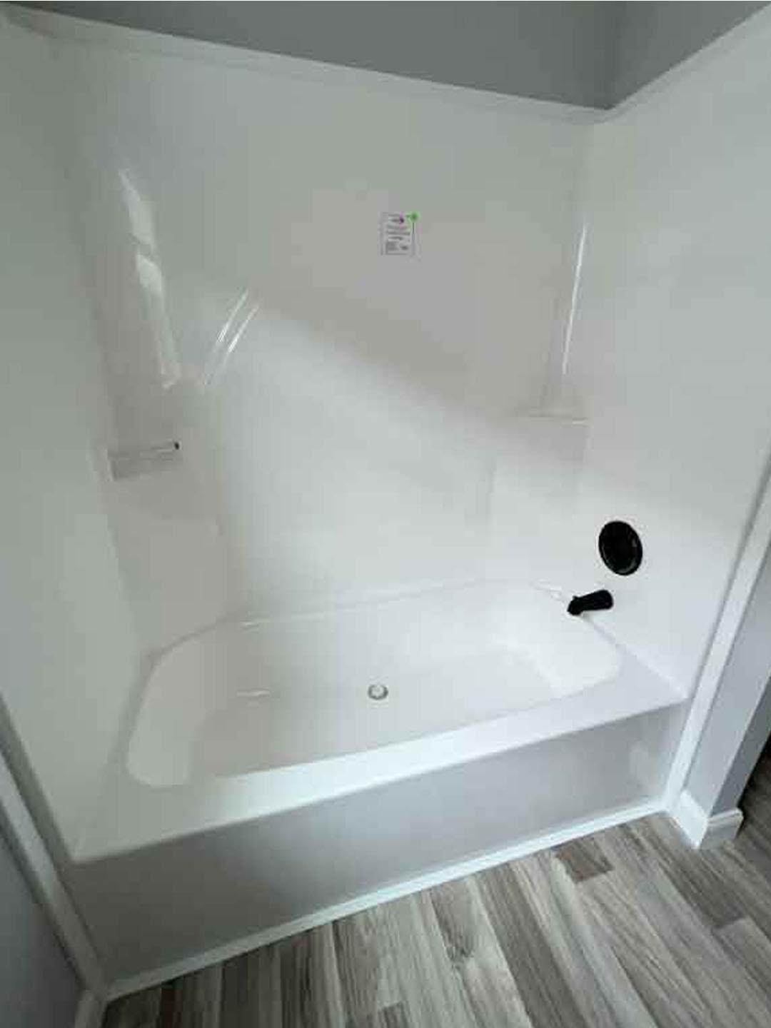 A clean white bathtub with a high-gloss finish is set against light gray walls and wood-patterned flooring. The faucet and drain are black.
